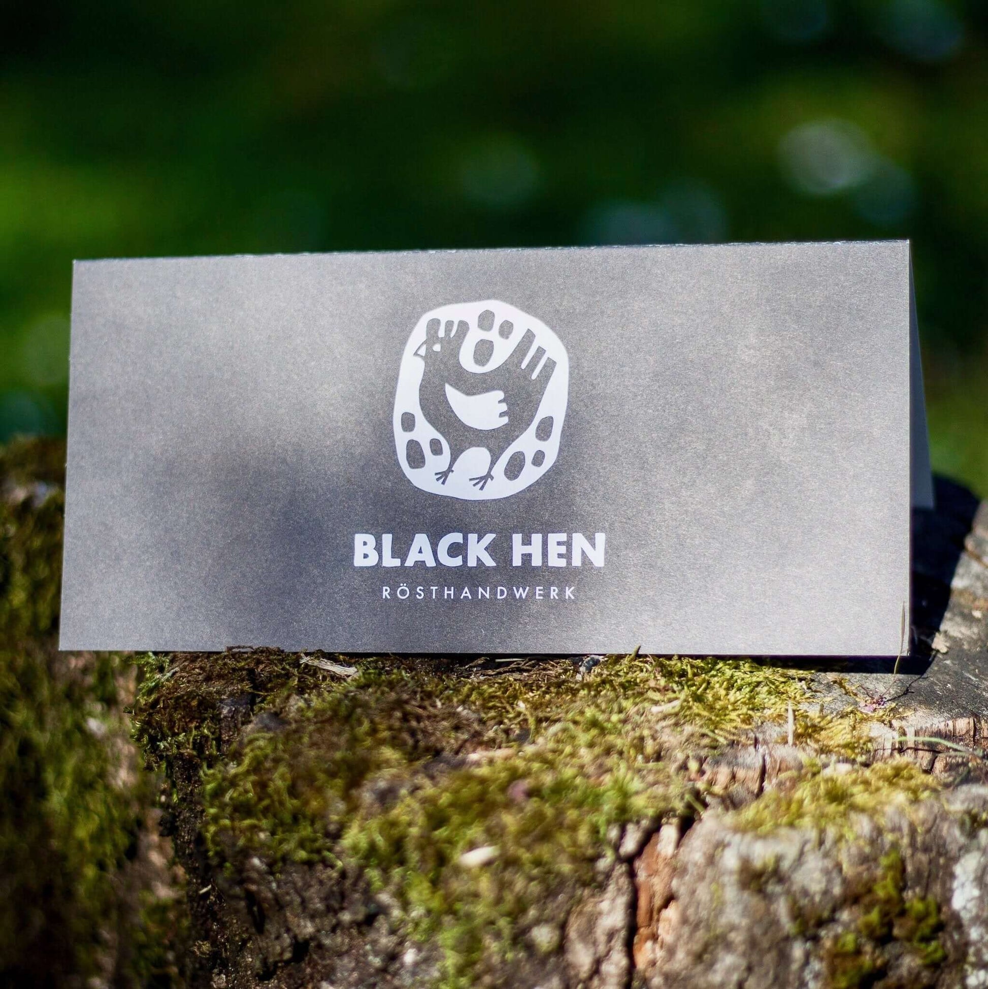 Black Hen logo on a gray card displayed on a moss-covered log outdoors.