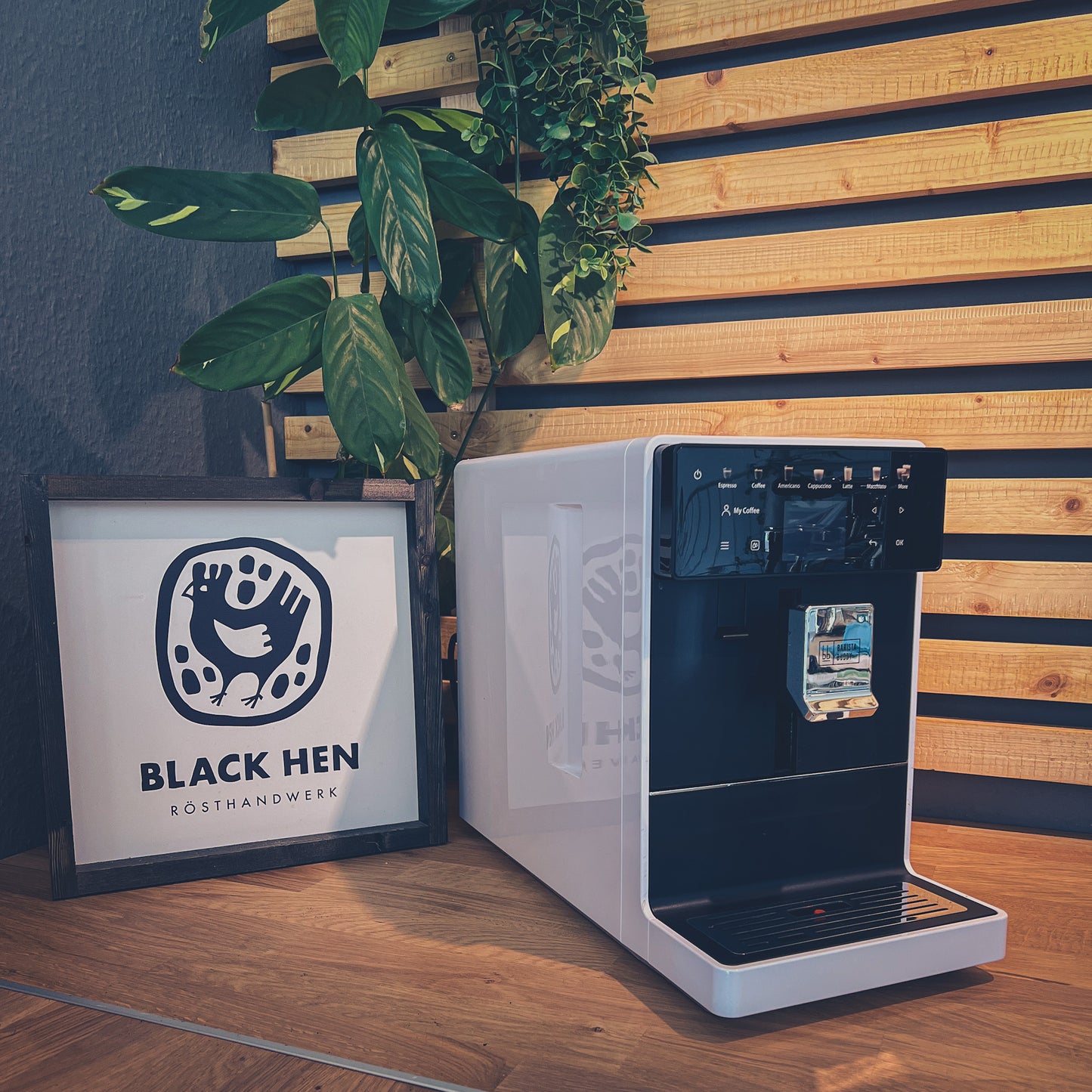 Gutschein Vollautomaten-Workshop "Push the Button" vor dem Black Hen-Schild mit Kaffeebohnen und Pflanzen im Hintergrund.