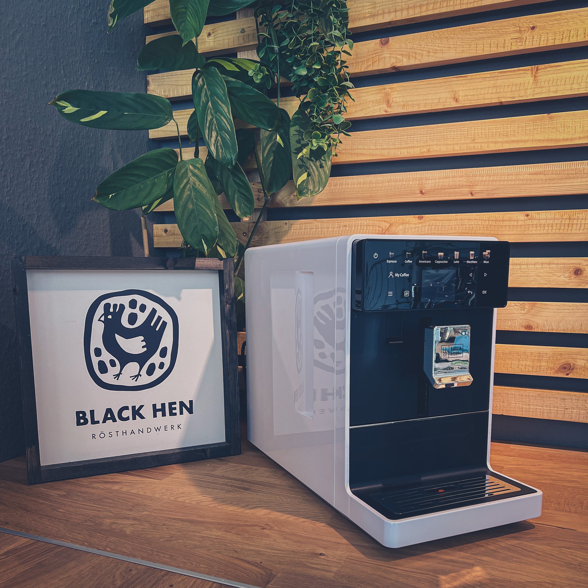 Gutschein Vollautomaten-Workshop "Push the Button" vor dem Black Hen-Schild mit Kaffeebohnen und Pflanzen im Hintergrund.