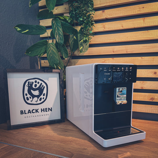 Gutschein Vollautomaten-Workshop "Push the Button" vor dem Black Hen-Schild mit Kaffeebohnen und Pflanzen im Hintergrund.